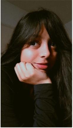 Headshot of young female, wearing a dark jumper, resting her chin on her hand as she gazes towards the lens. Her dark hair frames her face.