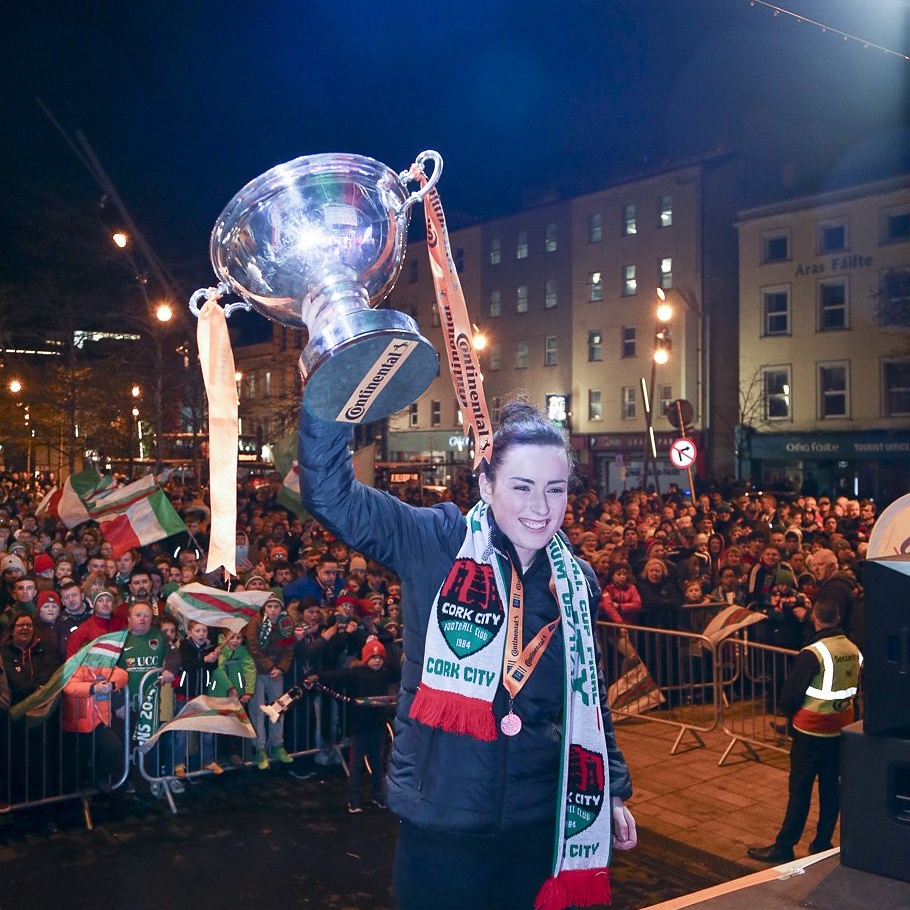Ciara McNamara, Captain, Cork City Football Club. Young girl with dark hair wearing a Cork City red, green and white scarf around her neck along with a winners medal. Her arm is raised triumphantly as she holds up a large silver cup. Behind her a large crowd is gathered.