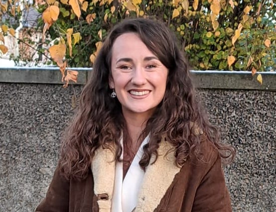 Head and shoulder shot of a smiing student with long dark hair, wearing a brown sheepskin coat, against a grey brick wall.