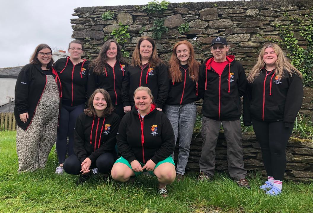 A group of nine students line up in two rows and pose for the camera in front of a brick wall flecked with green fern and ivy. They are all wearing black hoodies with red detail. The hoodies have the UCC logo stitched on the front.