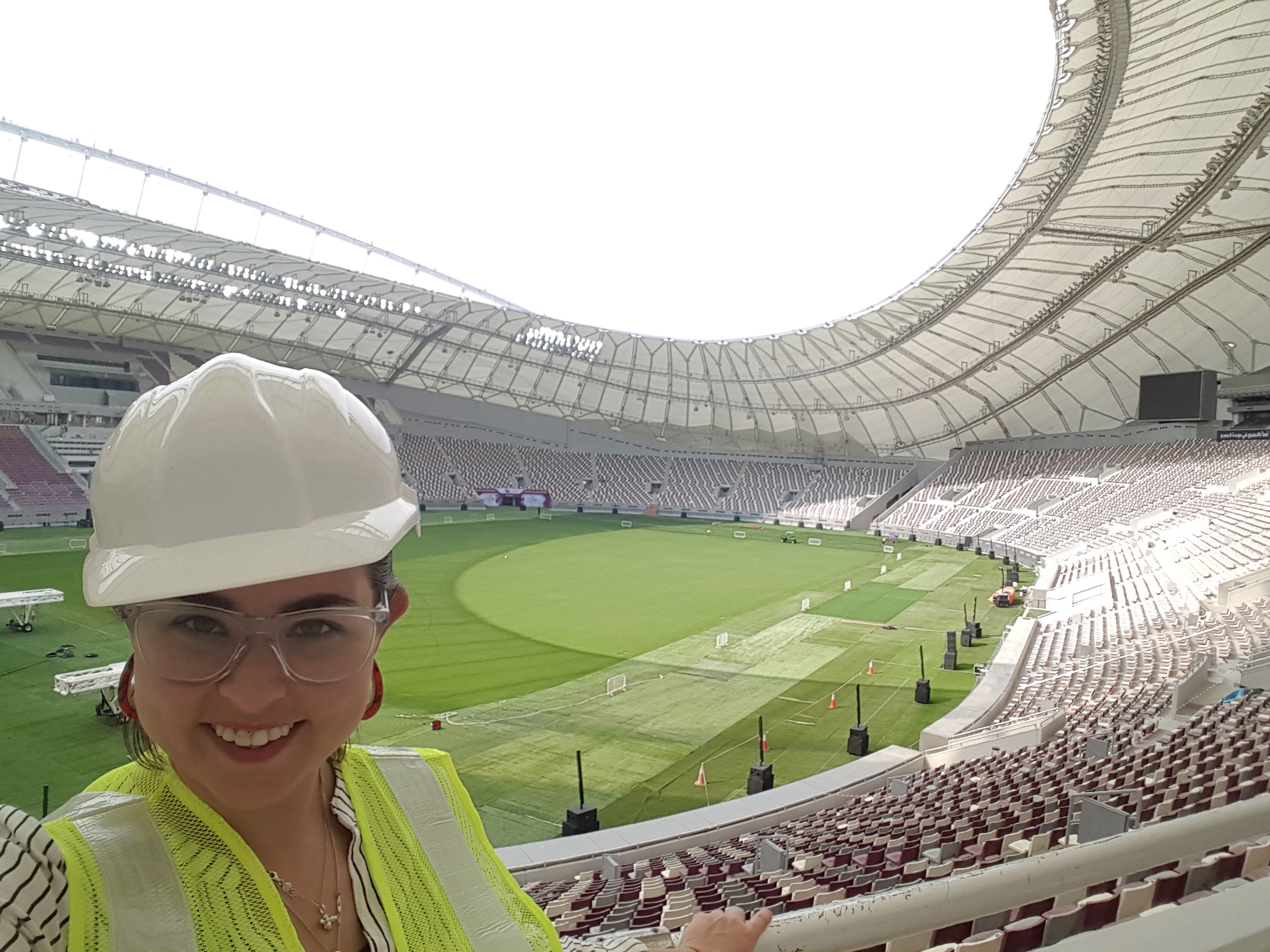 Headshot of Museum Studies student Victoria in a white hard hat and yellow high-viz jakcet, poses in a large stadium.