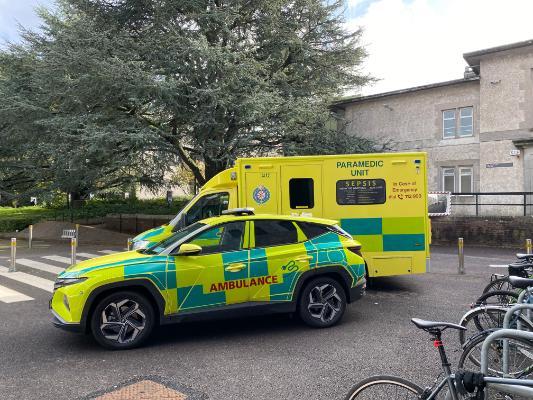 Yellow Ambulance and Yellow and Green responder car parked in the grounds of UCC against a backdrop of trees and pebble-dashed buildings.