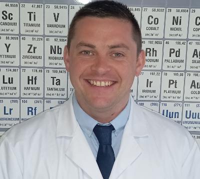 Headshot of ScEd graduate in a white coat, smiling for the camera, against a background displaying the periodic table.