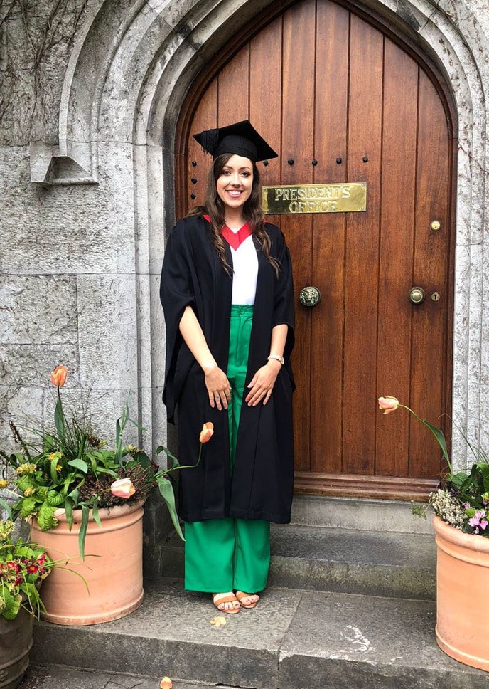 Student with long hair and wearing a graduation gown and flat graduation cap (mortarboard) poses in front of a brown door with a brass plaque engraved 'President's Office'.