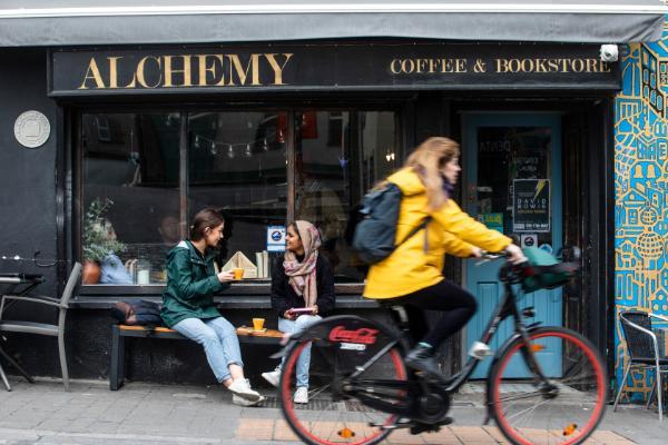 Female in yellow jacket on a bicycle cycling past the Alchemy coffee shop on Barrack Street in Cork. Two females are sitting outside the coffeeshop drinking coffee and chatting.