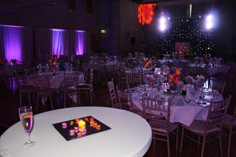 Foreground white table with a mirror centrepiece decorated in flower petals and a tea light. Background a beautifully decorated room with purple mood lighting.