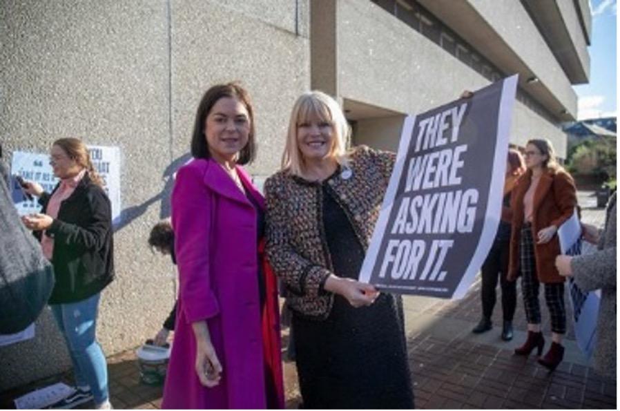 Photo of two UCC staff members standing in front of Bystander Intervention mural holding a sign saying 'They were asking for it'