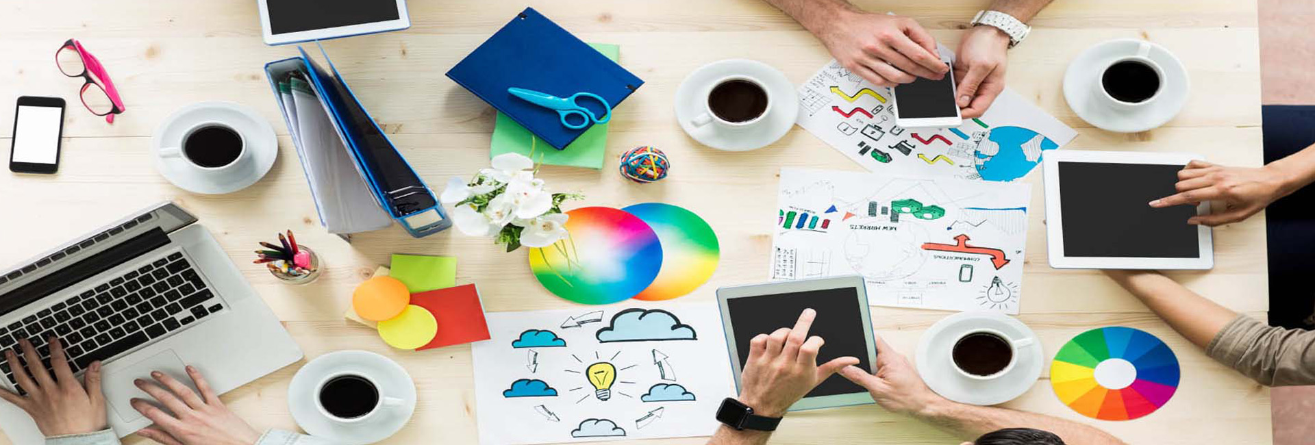 picture of a table with artwork, coffee, notebooks, and hands holding iPads