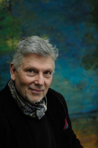 A white man with white hair in a dark top against a dark background looking at the camera.