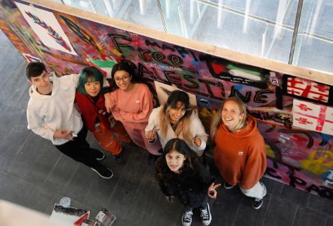 A group of young people looking up at the camera and standing in front of a large artwork.
