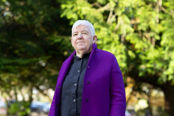 A woman with very short, white hair wearing a purple jacket looking at the camera with a half smile