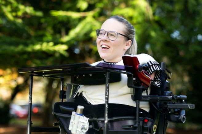A smiling woman wearing glasses looking off into the distance in a wheelchair