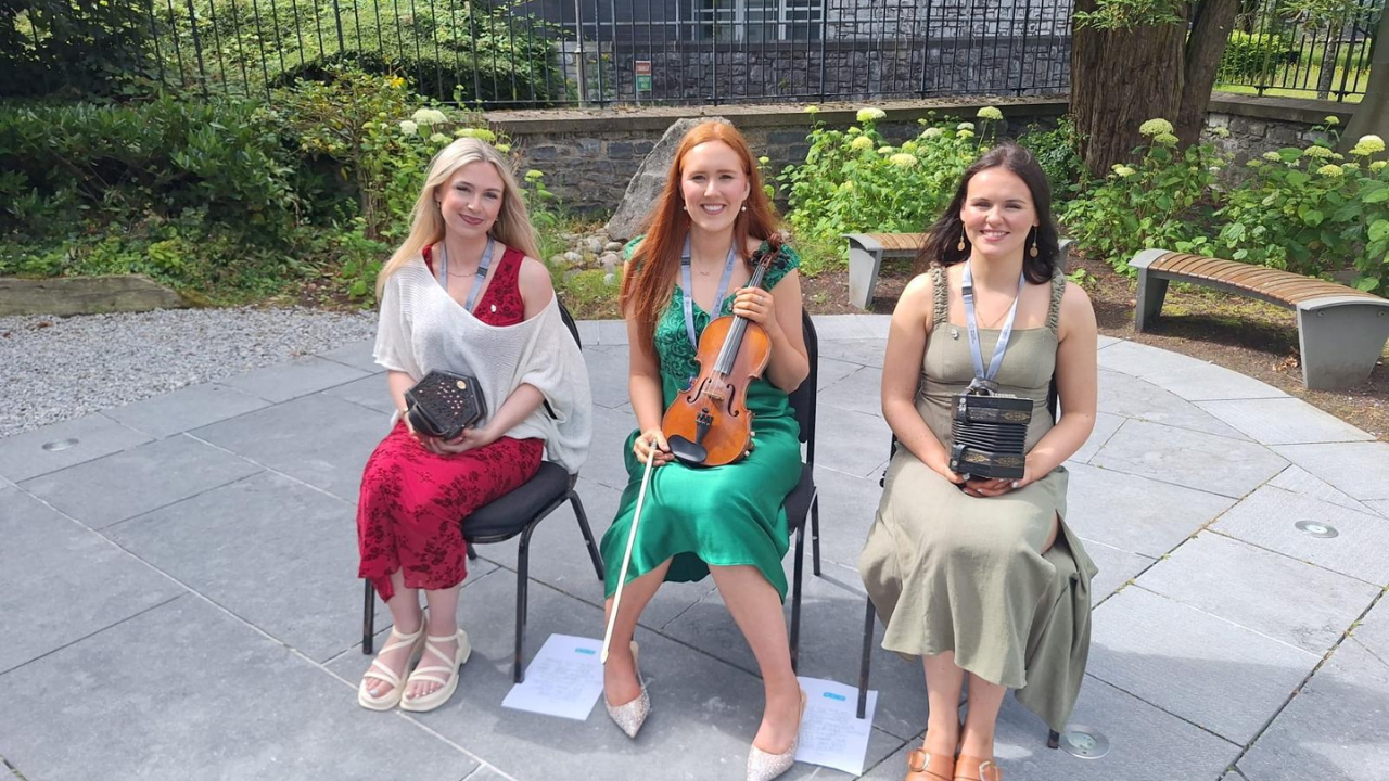 This is a picture of three Quercus Talented Students' Programme Creative & Performing Arts Scholars sitting down outside the Honan Chapel in UCC with their instrucments. Gráinne Ahern is on the left with a concertina, Eimear Kellerher is in the middle with a fiddle, & Ríona Hely is on the right with a concertina.