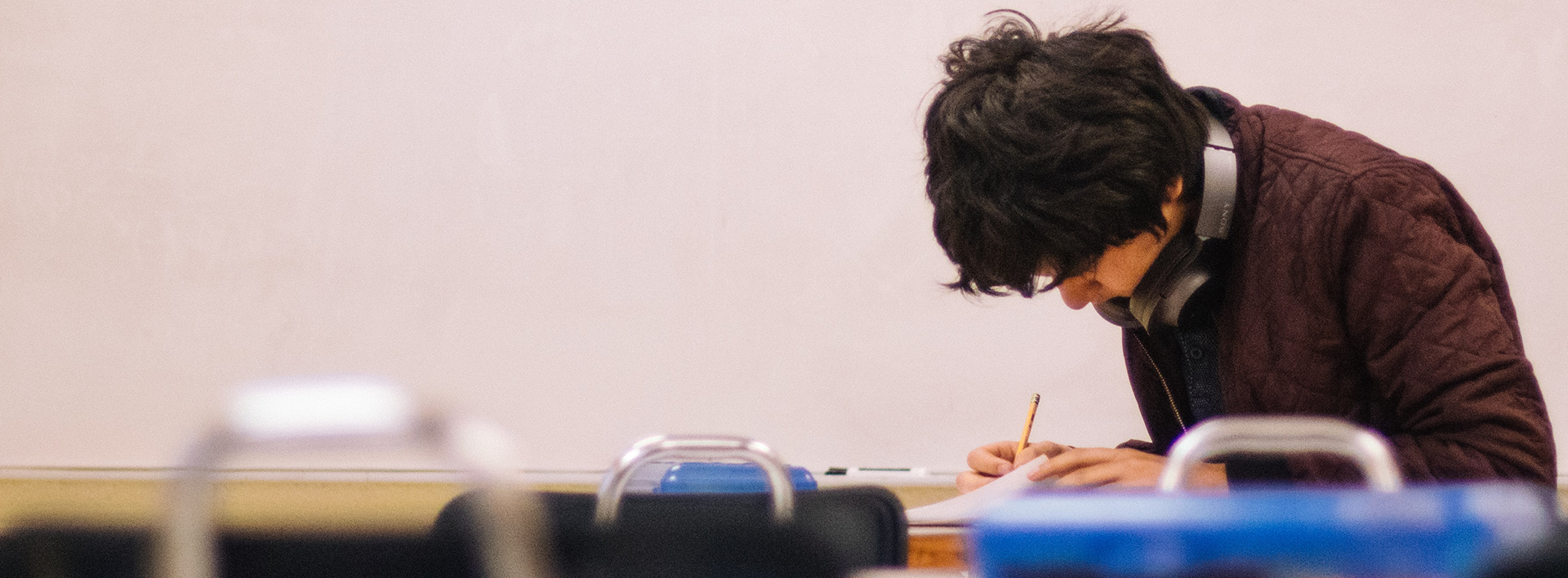 A student wearing a brown jacket sitting in a classroom writing.