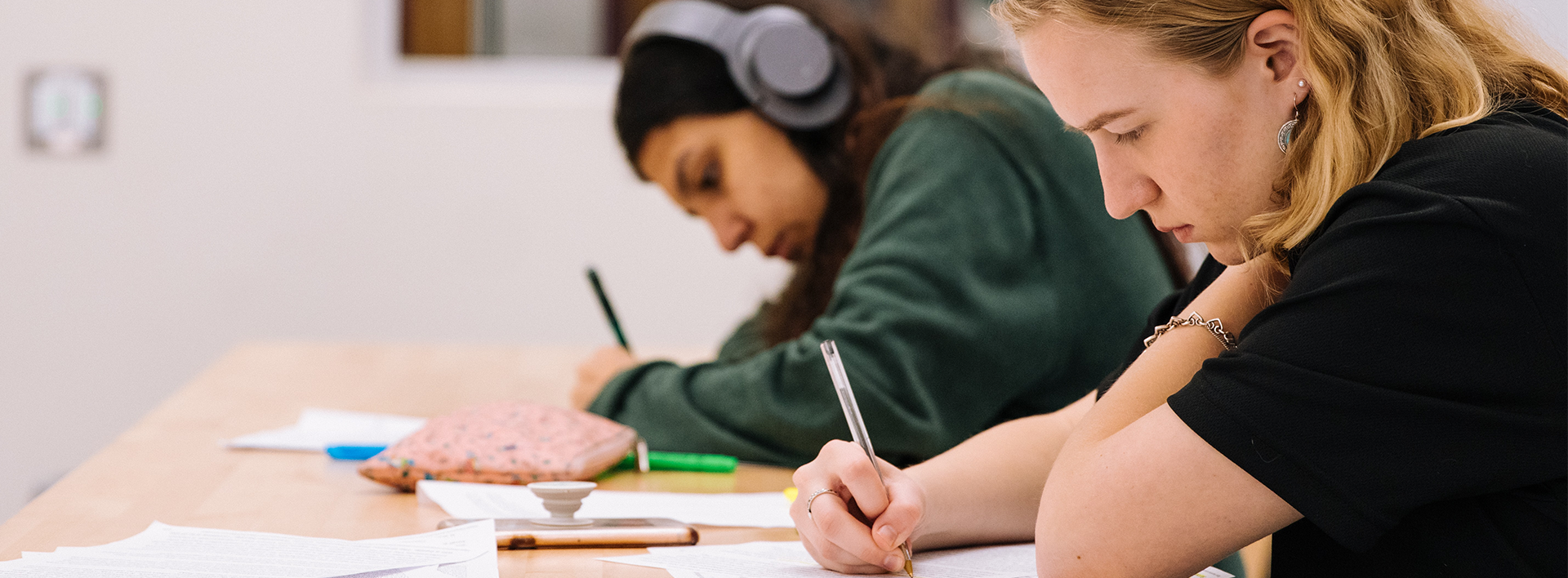 Two females students writing, one is wearing headphones.