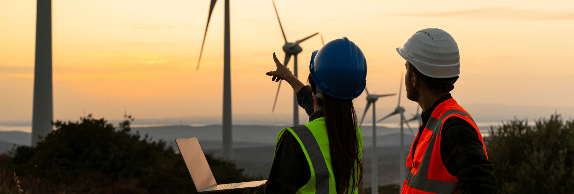 A man and a woman holding a laptop pointing towards wind turbines in background