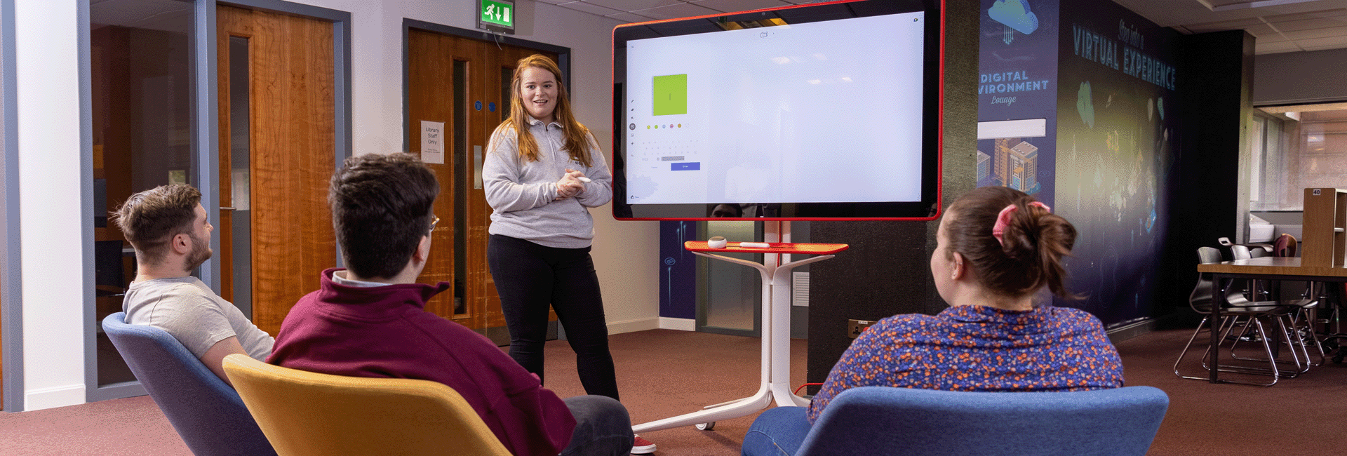 A young woman standing beside a large screen speaking to three young people sitting in front of her