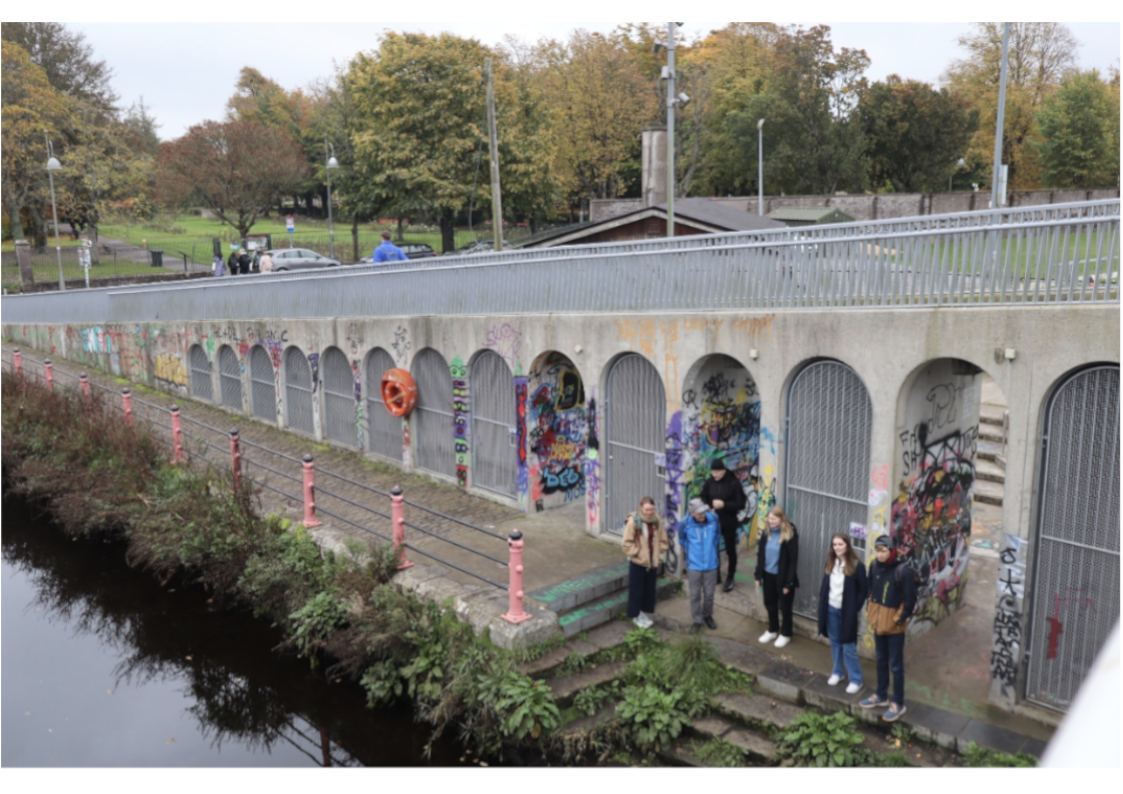 A group of students stand by the riverside featuring some old infrastructure