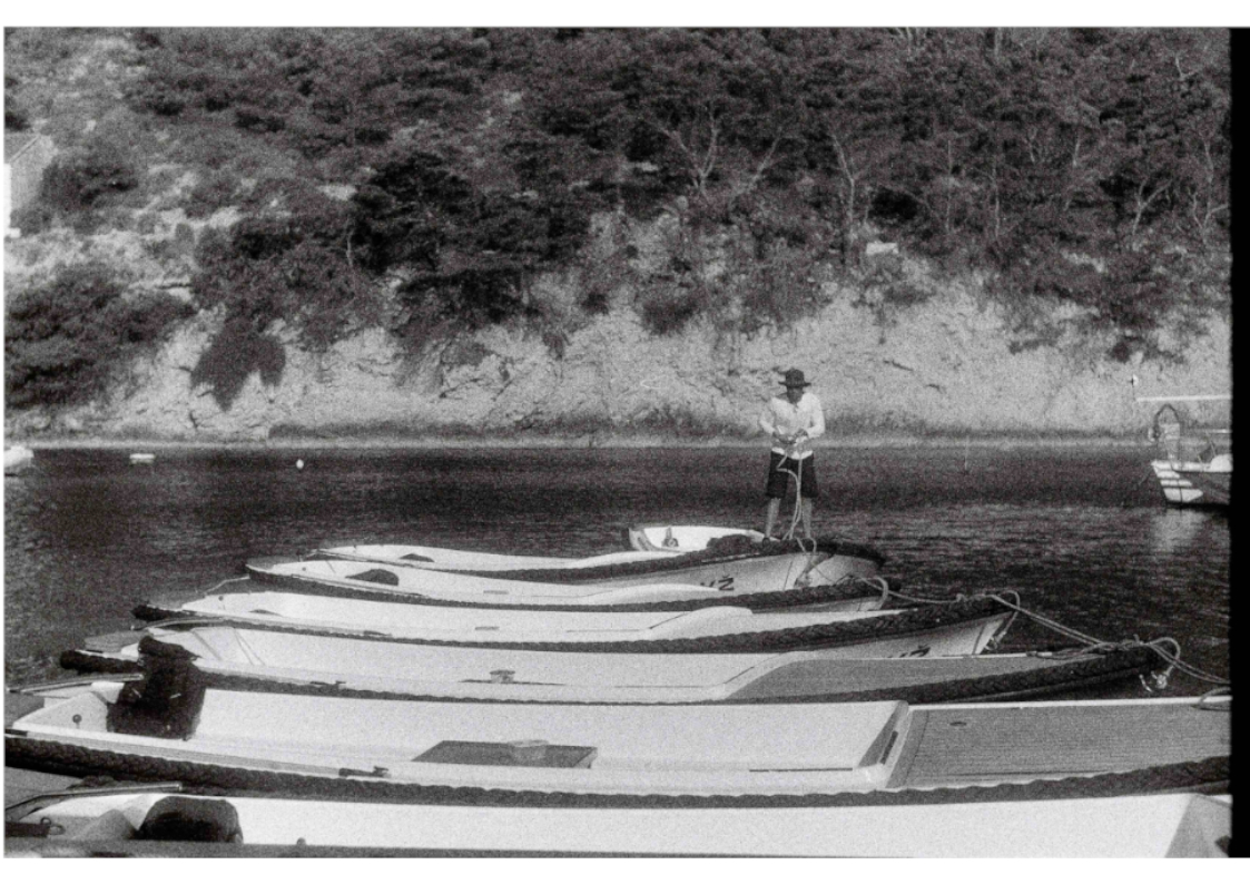 A man stands on the last of a row of traditional boats tied together, with the coastline behind.