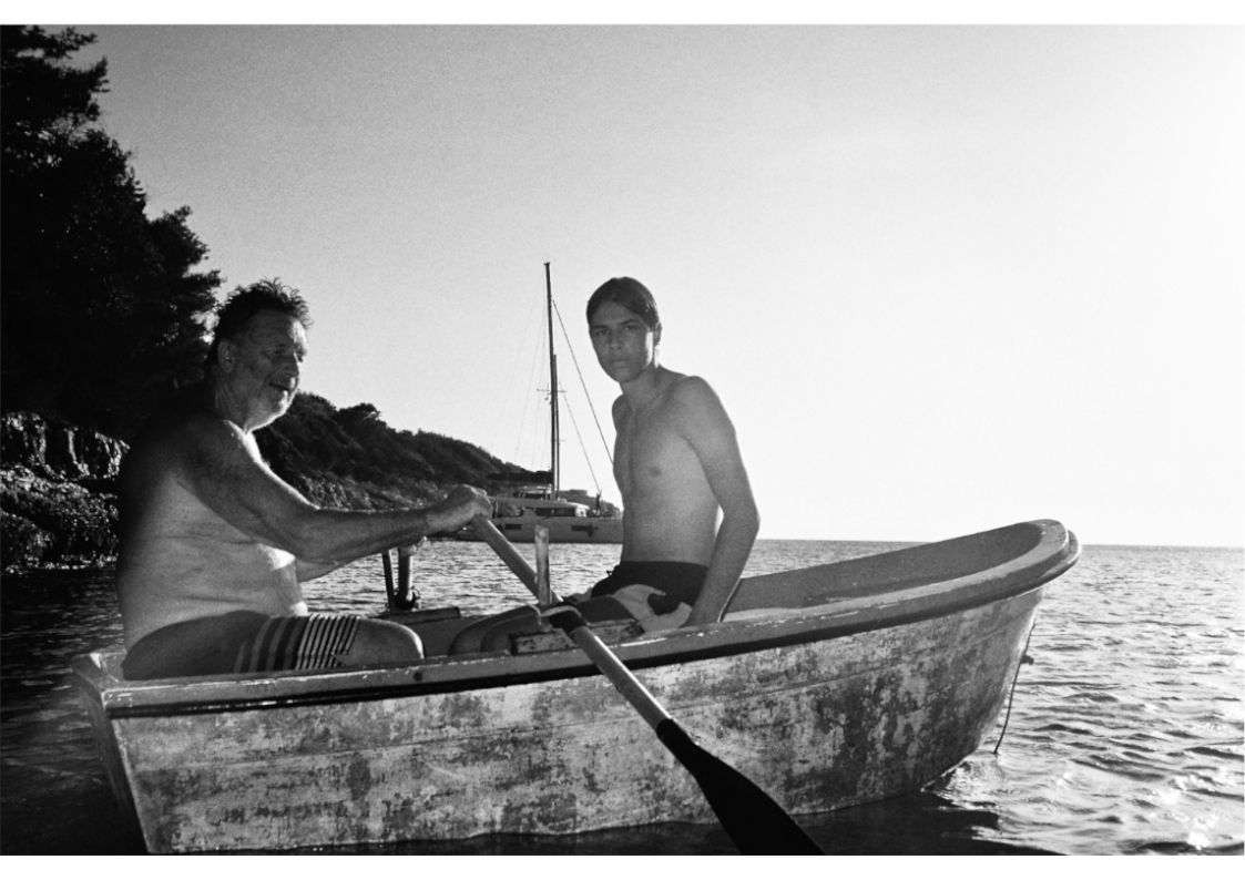 A young man and older man sit in a small traditional boat on the sea, in warm weather.